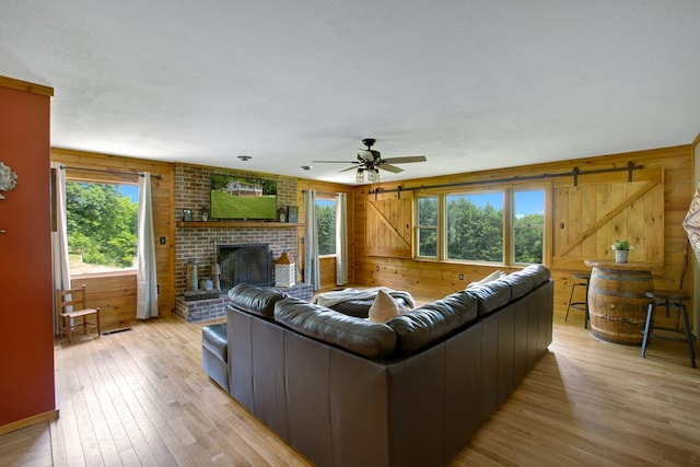 living area with a brick fireplace, light wood-style floors, wooden walls, a barn door, and a ceiling fan