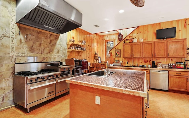 kitchen with a kitchen island with sink, appliances with stainless steel finishes, sink, wooden walls, and wall chimney exhaust hood