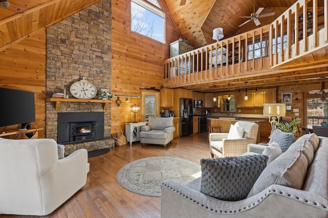 living room featuring wooden walls, a ceiling fan, a stone fireplace, wooden ceiling, and light wood-type flooring