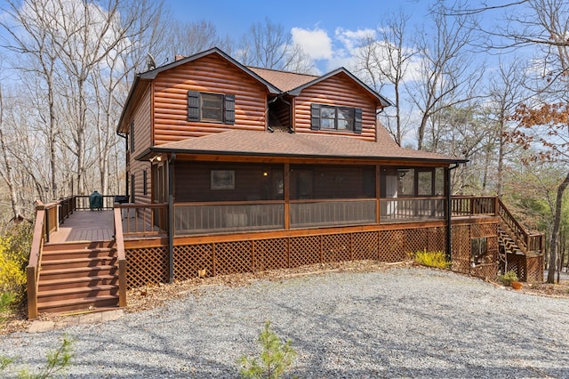 view of front facade featuring log veneer siding, roof with shingles, a sunroom, a wooden deck, and stairs