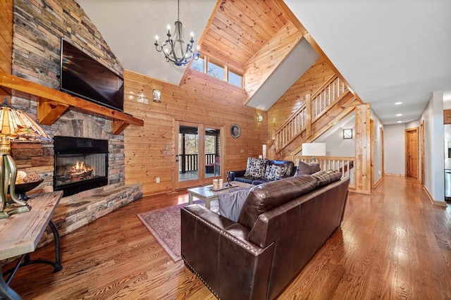 living room with wooden walls, wood-type flooring, high vaulted ceiling, a chandelier, and a stone fireplace