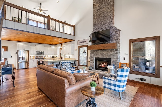 living area featuring high vaulted ceiling, light wood-type flooring, a ceiling fan, and a stone fireplace