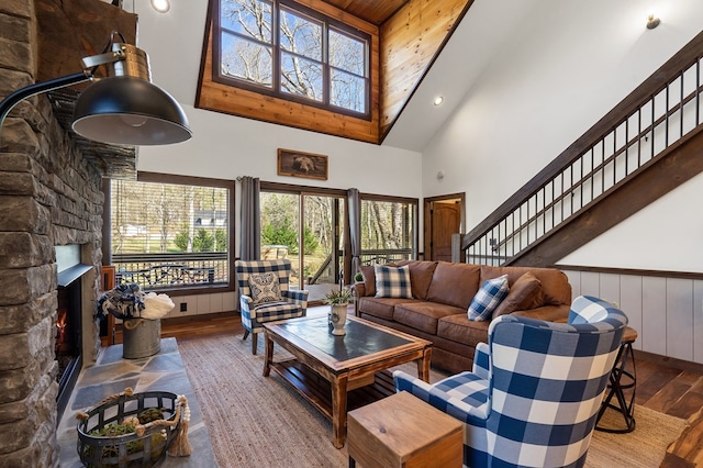 living area featuring high vaulted ceiling, wood finished floors, healthy amount of natural light, a stone fireplace, and wainscoting