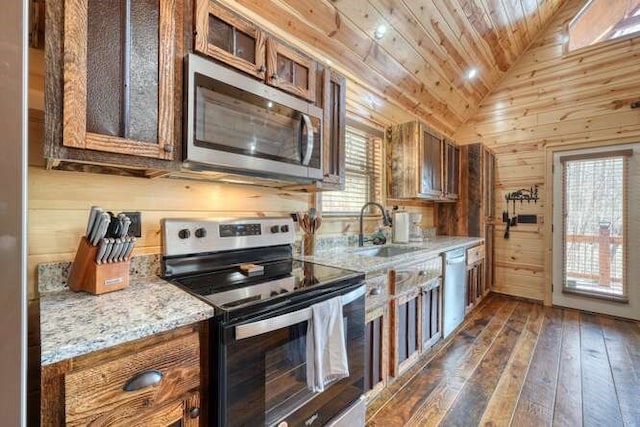 kitchen with wooden walls, lofted ceiling, appliances with stainless steel finishes, brown cabinetry, and a sink