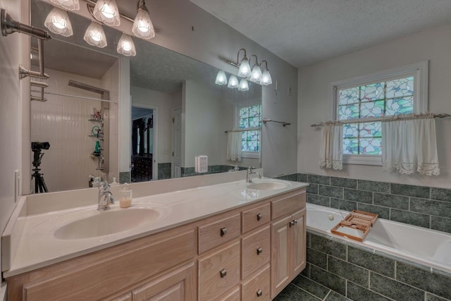 bathroom with a textured ceiling, a wealth of natural light, dual bowl vanity, and a relaxing tiled tub