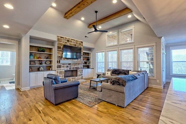 living room featuring ceiling fan, beam ceiling, light hardwood / wood-style floors, built in shelves, and a stone fireplace