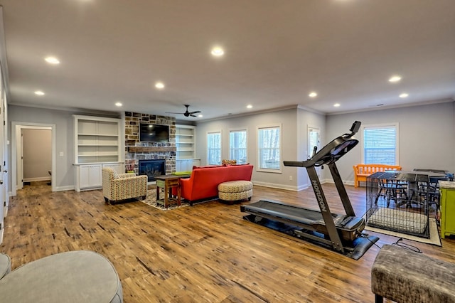 exercise area featuring hardwood / wood-style flooring, ceiling fan, ornamental molding, built in shelves, and a stone fireplace