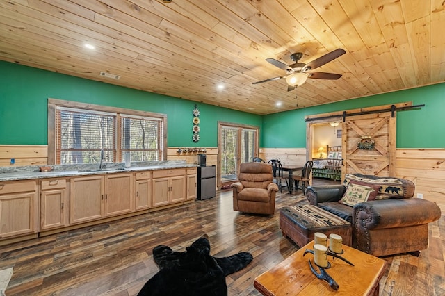 living room featuring a barn door, a ceiling fan, a wainscoted wall, wood ceiling, and dark wood-style flooring