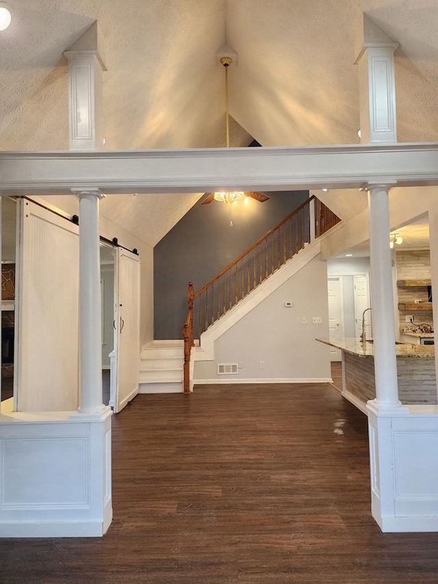 unfurnished living room with high vaulted ceiling, decorative columns, ceiling fan, a barn door, and dark wood-type flooring