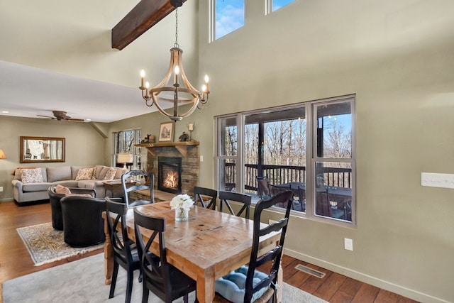 dining space featuring wood-type flooring, ceiling fan with notable chandelier, a stone fireplace, and a high ceiling