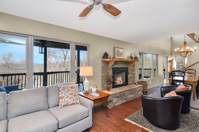 living room featuring a stone fireplace, dark wood-type flooring, a healthy amount of sunlight, and ceiling fan with notable chandelier