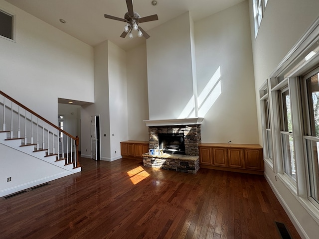 unfurnished living room featuring ceiling fan, a stone fireplace, baseboards, stairway, and dark wood finished floors