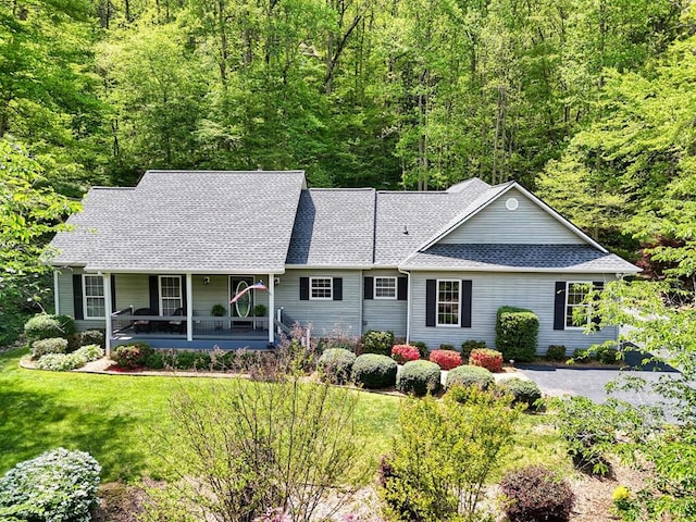 ranch-style home with roof with shingles, a forest view, a porch, and a front yard