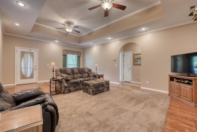living area with ceiling fan, arched walkways, ornamental molding, a tray ceiling, and light wood-style flooring