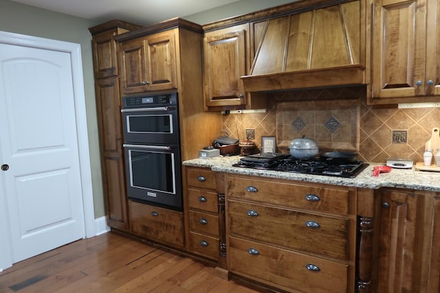 kitchen with custom exhaust hood, black gas stovetop, dark hardwood / wood-style floors, light stone countertops, and double oven