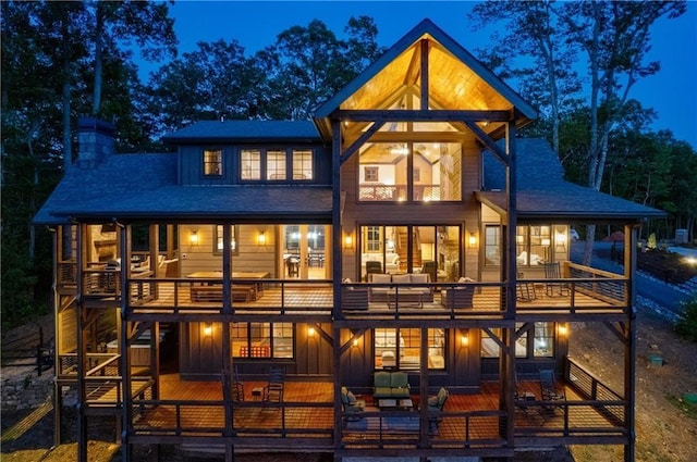 back of house at twilight featuring a balcony, a shingled roof, a chimney, and board and batten siding