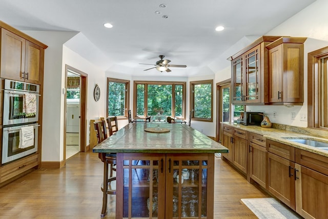 kitchen with light wood-type flooring, double oven, a kitchen breakfast bar, ceiling fan, and light stone counters