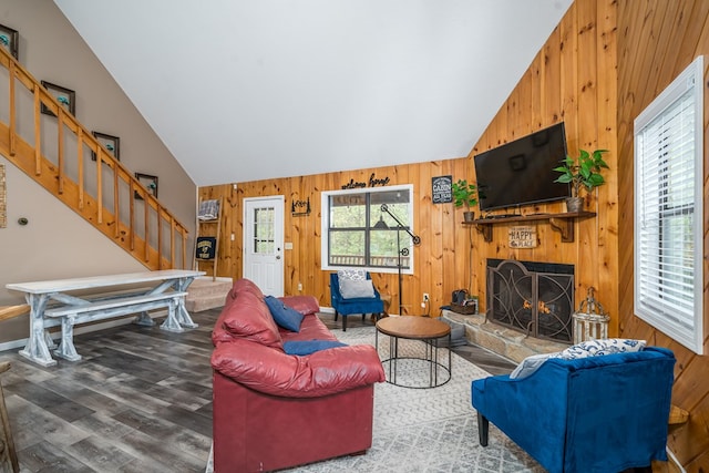 living area with high vaulted ceiling, stairs, wood walls, a lit fireplace, and wood finished floors