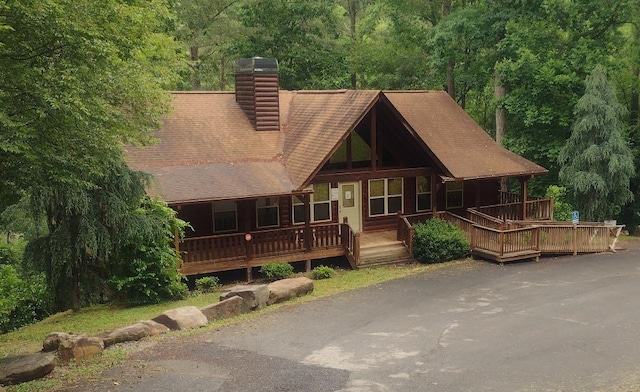 view of front facade with a shingled roof, covered porch, a chimney, and a view of trees