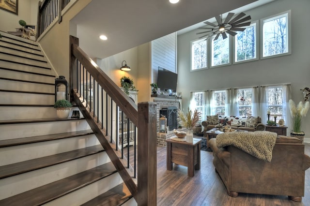 living room featuring dark wood-type flooring, ceiling fan, a brick fireplace, and a towering ceiling