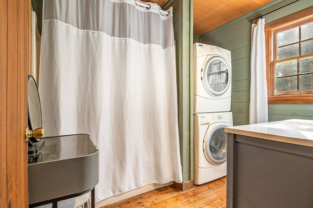 clothes washing area with wooden ceiling, sink, stacked washer and dryer, wooden walls, and light wood-type flooring