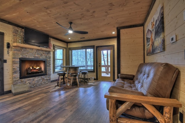 living area featuring ceiling fan, wooden ceiling, hardwood / wood-style floors, a stone fireplace, and wood walls