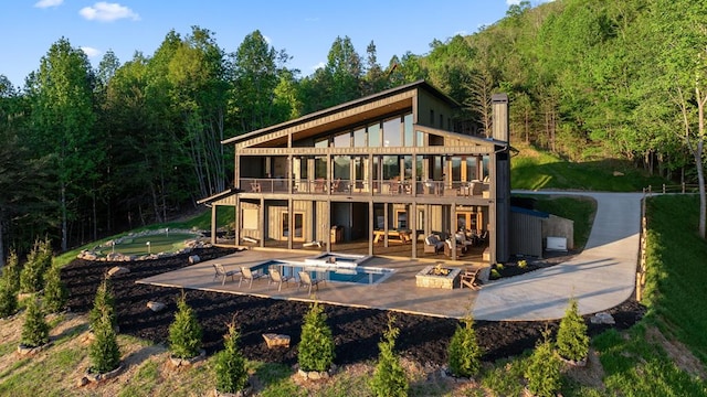 back of house featuring a forest view, a wooden deck, an outdoor fire pit, an outdoor hot tub, and a patio area