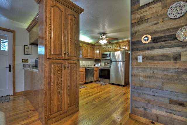 kitchen with wooden walls, a ceiling fan, light wood-style flooring, brown cabinetry, and appliances with stainless steel finishes