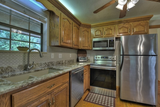 kitchen with a sink, ceiling fan, light stone countertops, brown cabinetry, and appliances with stainless steel finishes