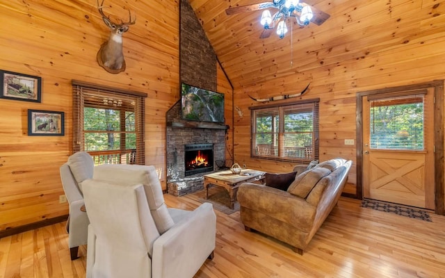 living room featuring high vaulted ceiling, a stone fireplace, wooden walls, ceiling fan, and light wood-type flooring