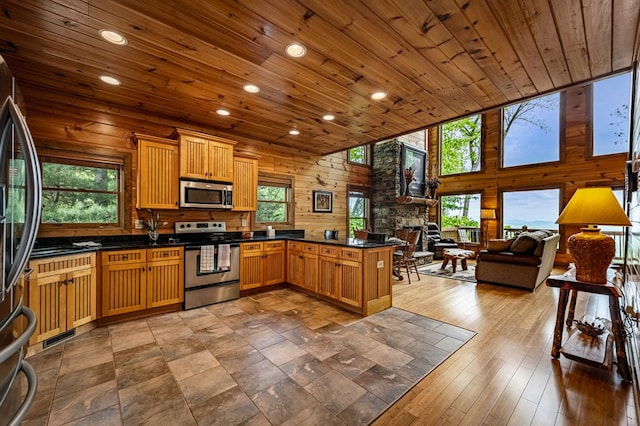 kitchen with a stone fireplace, appliances with stainless steel finishes, dark countertops, wooden ceiling, and a peninsula