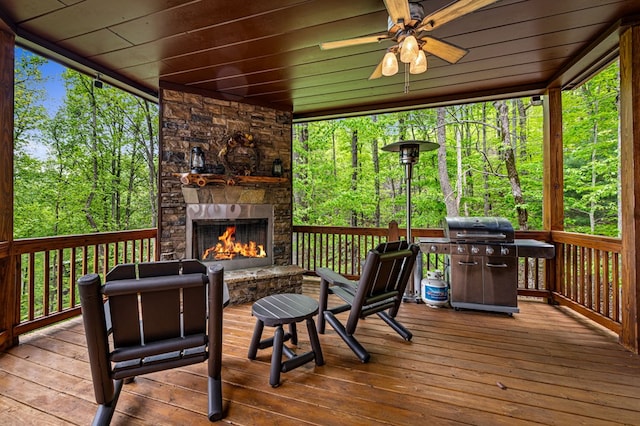 wooden deck featuring area for grilling, an outdoor stone fireplace, ceiling fan, and a forest view