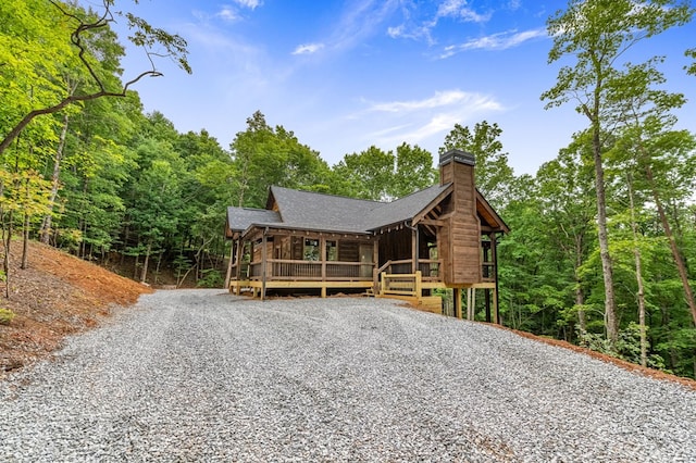 view of front of house featuring gravel driveway, a wooded view, a chimney, and a shingled roof