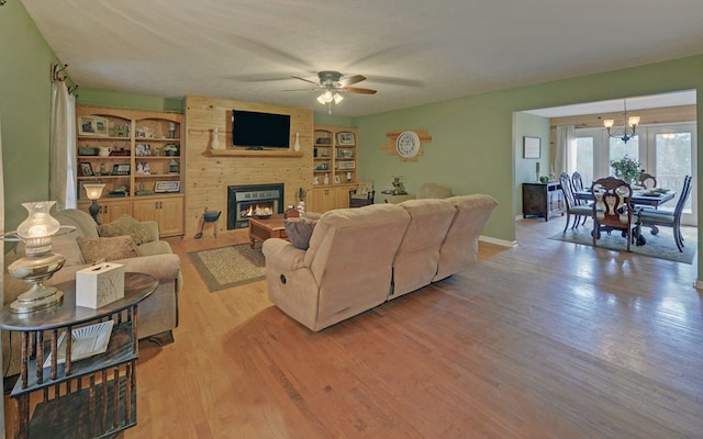 living room featuring built in shelves, a large fireplace, ceiling fan with notable chandelier, and light hardwood / wood-style flooring