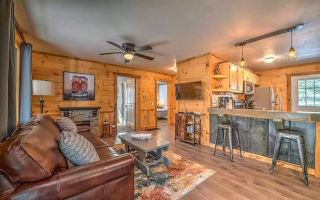 living room with ceiling fan, sink, light hardwood / wood-style floors, a stone fireplace, and wood walls