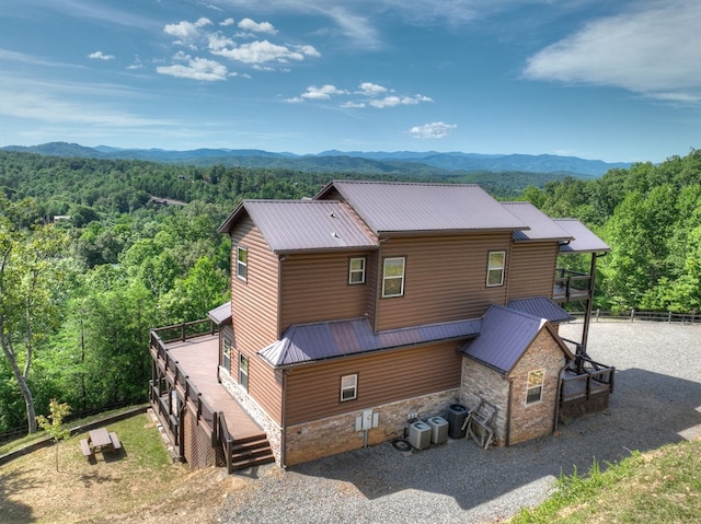 back of house featuring metal roof, central AC unit, stone siding, driveway, and a wooded view