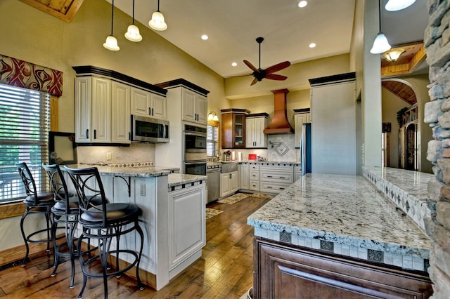 kitchen with dark wood finished floors, stainless steel appliances, custom range hood, light stone countertops, and a peninsula