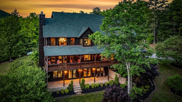 back of property at dusk featuring a chimney, a patio area, roof with shingles, and a deck