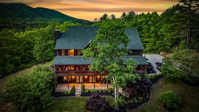back of property with a chimney, a patio, a view of trees, roof with shingles, and a deck