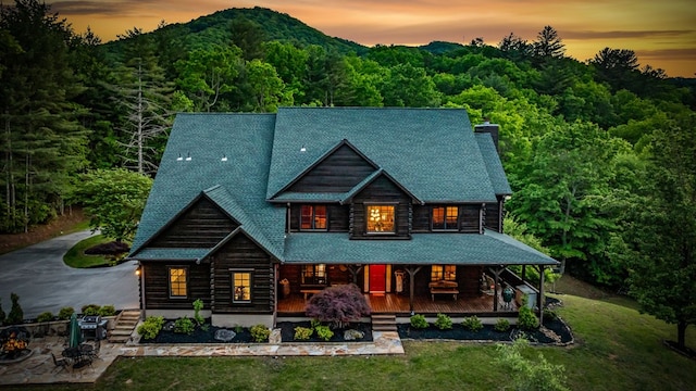 back of property at dusk featuring covered porch, a wooded view, a yard, log exterior, and a chimney