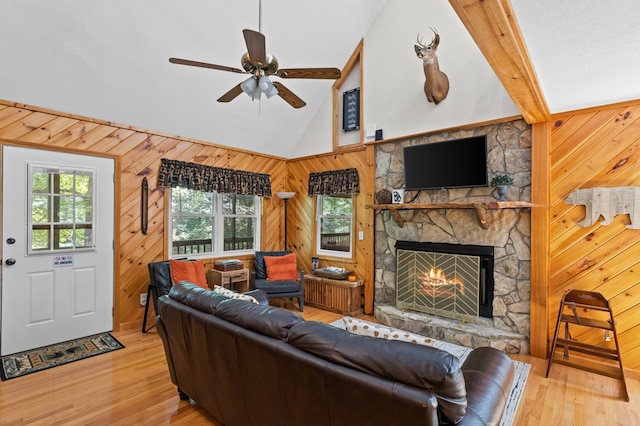 living room with light hardwood / wood-style flooring, ceiling fan, lofted ceiling with beams, a stone fireplace, and wood walls