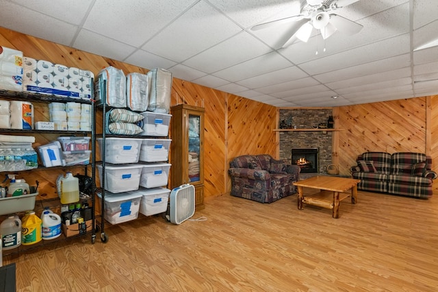 living room with a stone fireplace, a paneled ceiling, wood walls, ceiling fan, and light wood-type flooring