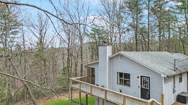 view of side of property with a deck, a wooded view, roof with shingles, and a chimney