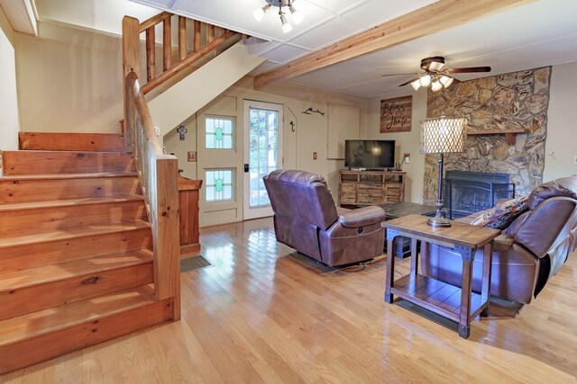 living room featuring light hardwood / wood-style floors, a stone fireplace, beam ceiling, and ceiling fan