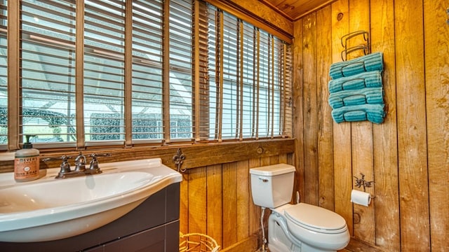 half bath with toilet, vanity, wood walls, and a healthy amount of sunlight