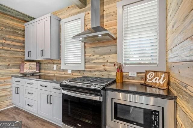 kitchen with hardwood / wood-style floors, white cabinets, wall chimney range hood, wooden walls, and appliances with stainless steel finishes