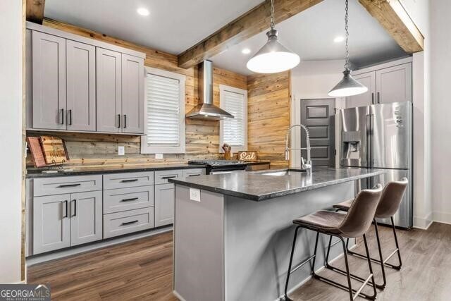 kitchen featuring a kitchen island with sink, wall chimney range hood, hanging light fixtures, sink, and stainless steel refrigerator with ice dispenser