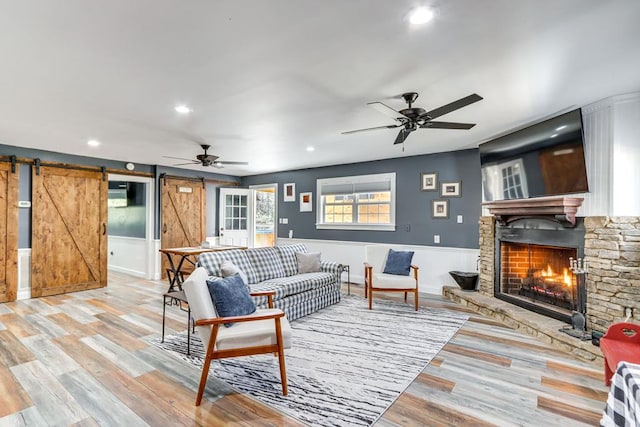 living room featuring a stone fireplace, light hardwood / wood-style floors, a barn door, and ceiling fan