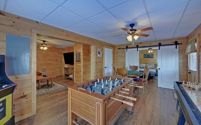 playroom with wooden walls, wood finished floors, electric panel, ceiling fan, and a barn door