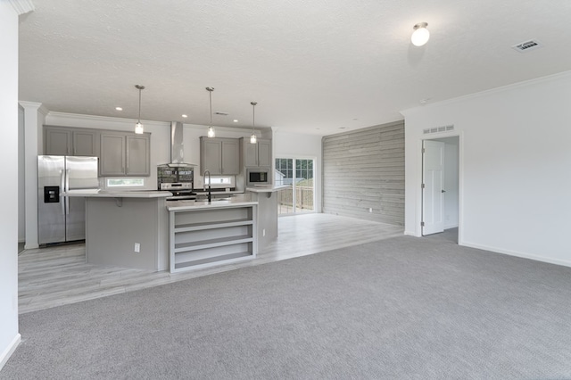 kitchen featuring hanging light fixtures, stainless steel appliances, wall chimney range hood, gray cabinets, and a center island with sink